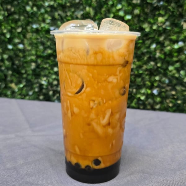 Iced Thai tea with boba and ice in a clear plastic cup against a green leafy background.
